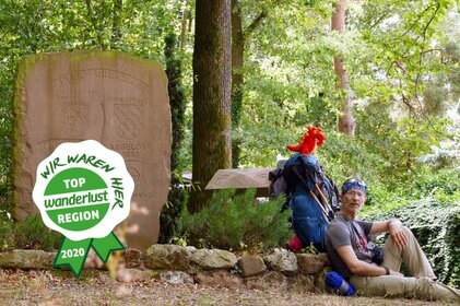 Fuat Gören sitzt im Wald neben einem Grenzstein auf dem Boden mit seinem Rucksack und dem roten Kuscheldrachen Nila. Auf dem Bild ist ein grün-weißes Logo mit dem Schriftzug "Top Wanderlust Region 2020, wir waren hier"