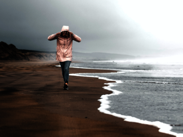 Person mit Regenjacke gekleidet läuft bei regnerischem Wetter an der Küste entlang durch den Sand