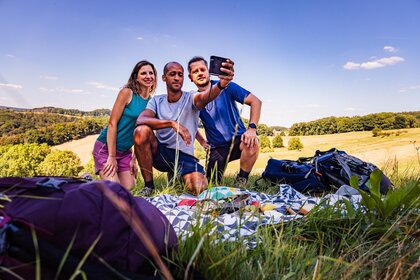 Drei Personen knien auf einer Picknickdecke auf einer grünen Wiese und machen ein Selfie mit einer Sofortbildkamera.