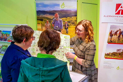 Am Messestand zeigt Mareike Müller zwei interessierten Besuchern eine Karte. Im Hintergrund ist ein Plakat des Odenwaldes mit Wanderern in der Natur. Rechts im Bild ist ein Plakat des Alemannenwegs mit verschiedenen Bildern.